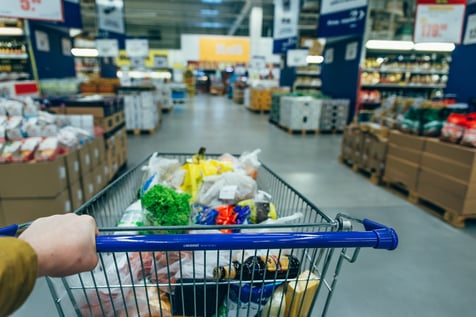 A first-person perspective of a shopper's hand pushing a metal shopping cart filled with groceries, including fresh produce and packaged goods, down a wide aisle in a brightly lit supermarket.