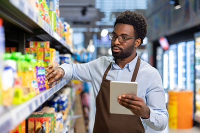 A grocery store employee wearing an apron stands in an aisle holding a digital tablet to check inventory data while organizing packaged goods on the supermarket shelves.
