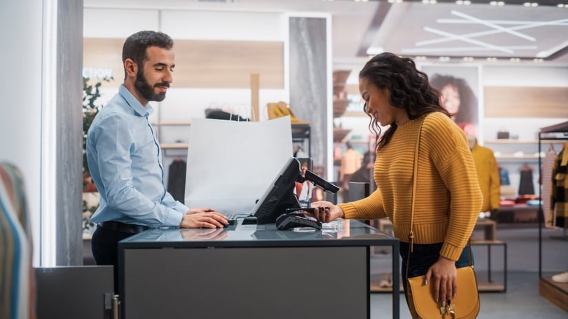 Young Woman at Counter Buys Clothes from Friendly Retail Sales Assistant, Paying with Contactless NFC Smartphone Touching Terminal