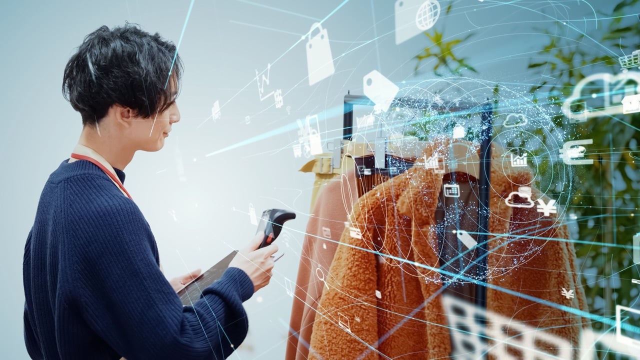 A retail employee uses a handheld RFID scanner to check a rack of coats. A digital network graphic overlays the clothing, featuring interconnected icons of price tags, shopping bags, and data clouds to represent wireless inventory tracking.
