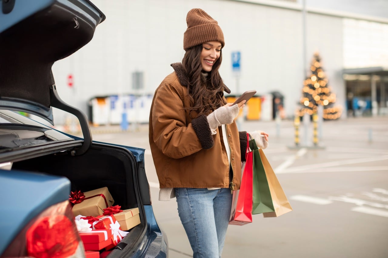 a young woman in winter jacket and stocking cap holding shopping bags and cellphone in standing near car outside a retail shopping mall with holiday tree in background.
