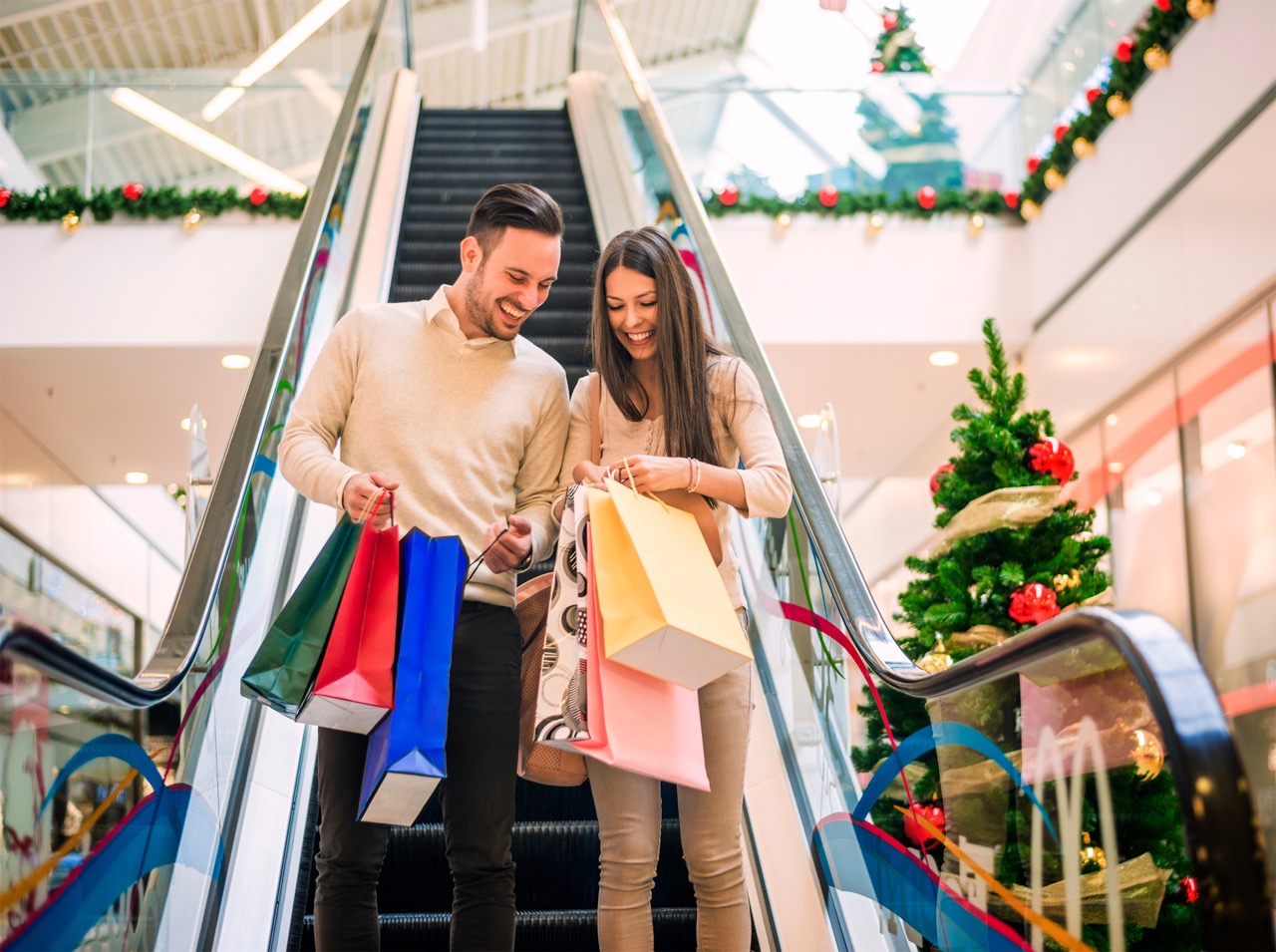 A smiling man and woman ride down a shopping mall escalator, looking happily at the multiple brightly colored shopping bags they are holding. The mall is decorated for the holidays with garland and a Christmas tree in the background.