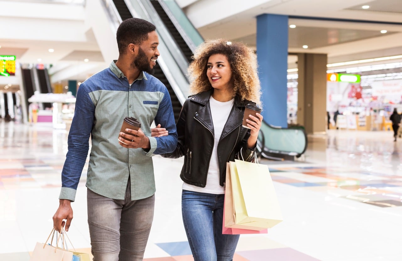 two young adults walking arm in arm in shopping mall holding shopping bags and coffee cups