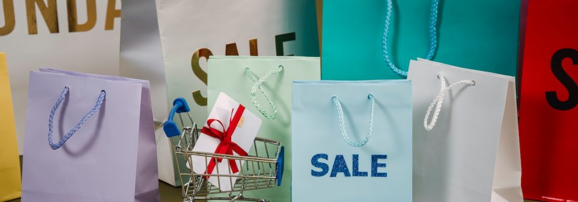 A blue shopping bag with "sale" on the front, and a small shopping cart with a gift card inside. Behind them are a variety of shopping bags.